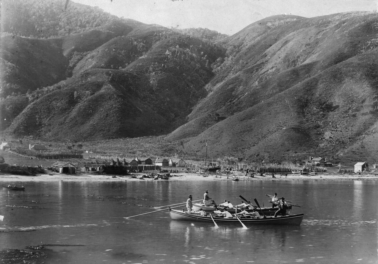 Two whaleboats with crews and harpoons on the water in front of a small beach settlement